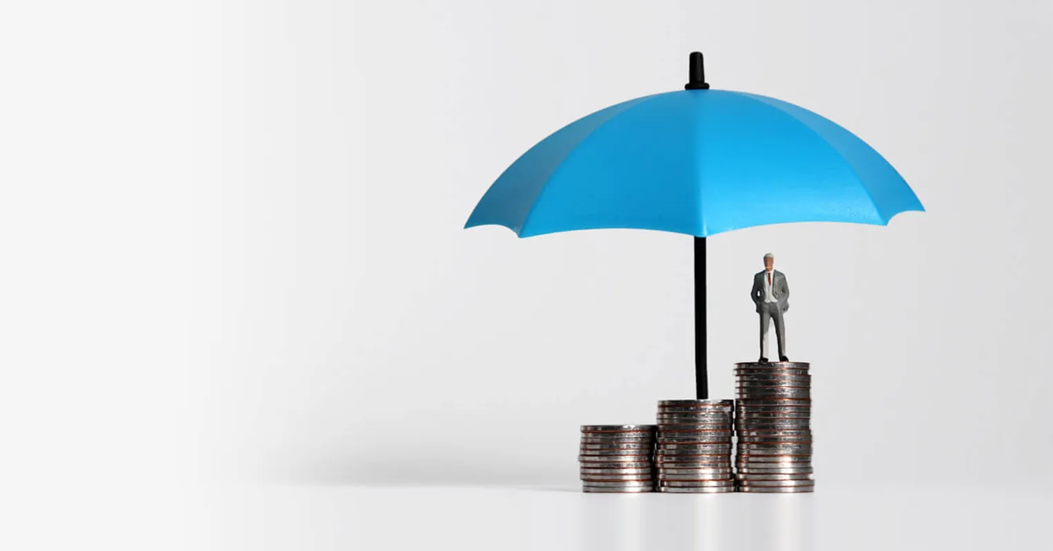 Three different sized stacks of coins, with a toy figure of a man stood on the tallest, with a blue umbrella sheltering them all
