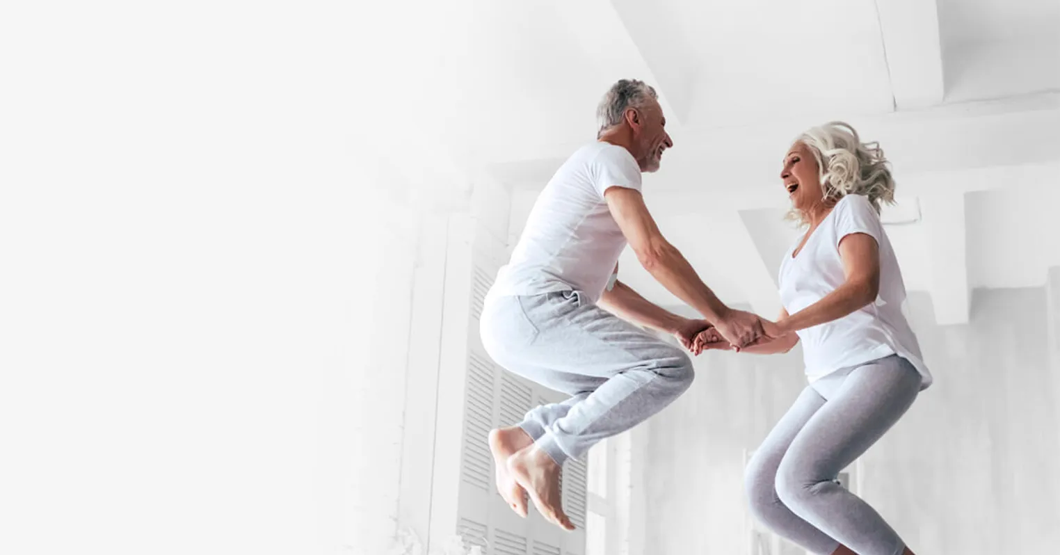 Photo of a man and woman wearing grey jogging pants and white t-shirts jumping up into the air and laughing